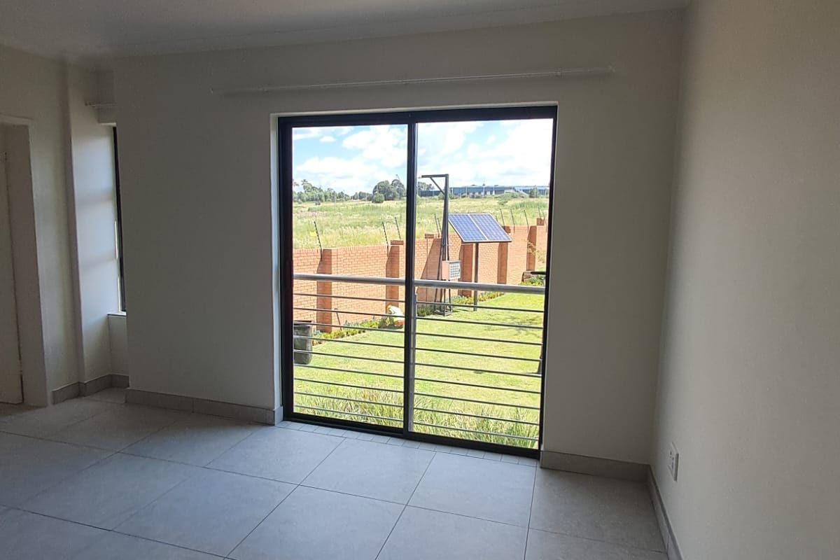 A bright, empty living room featuring neutral tiled flooring and a large sliding glass door with security bars, offering views of the garden.