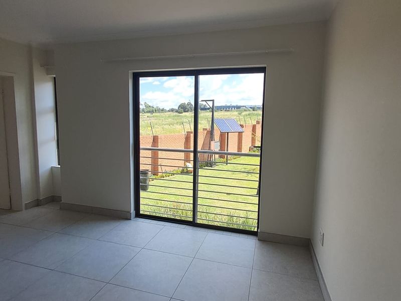 A bright, empty living room featuring neutral tiled flooring and a large sliding glass door with security bars, offering views of the garden.