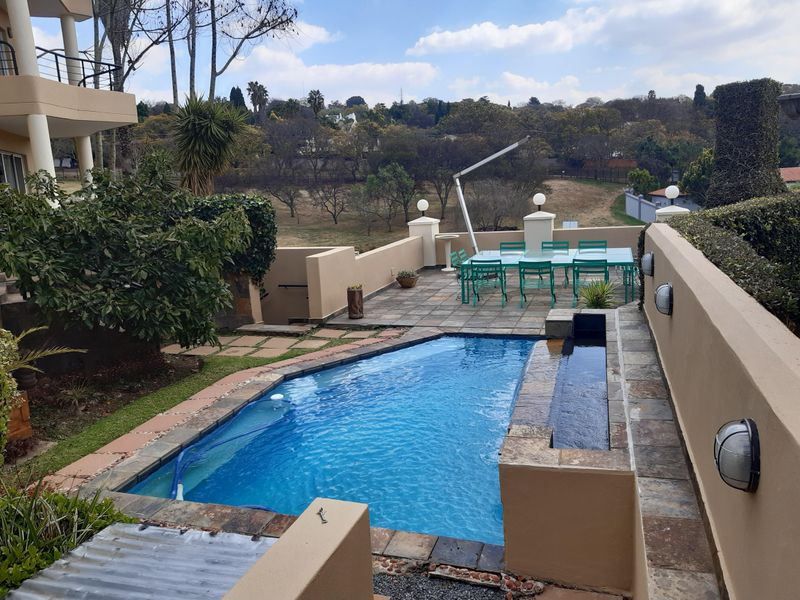 An elevated outdoor pool area featuring a tiled patio, built-in water feature, and retaining walls, overlooking a landscaped garden and surrounding estate.