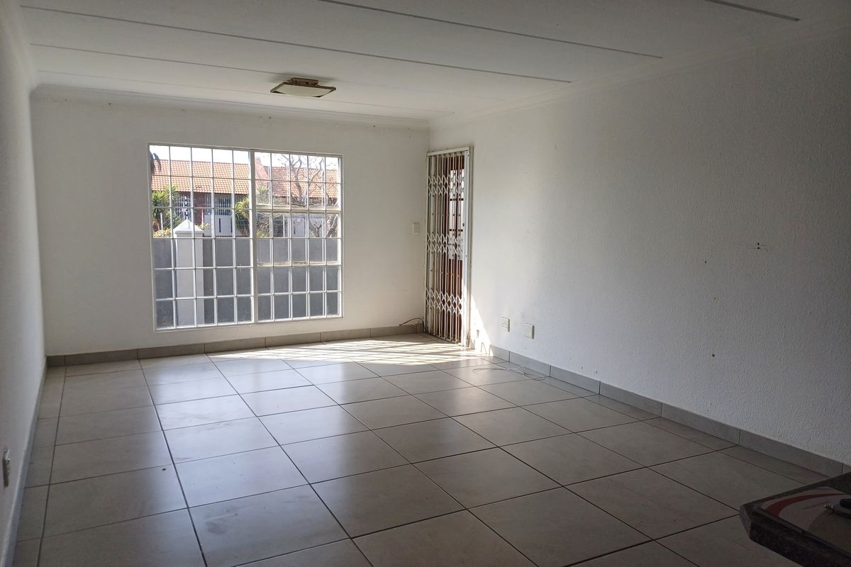 An empty, open-plan living area featuring neutral tiled flooring, a security gate, and large windows with burglar bars, offering a blank canvas for interior design.