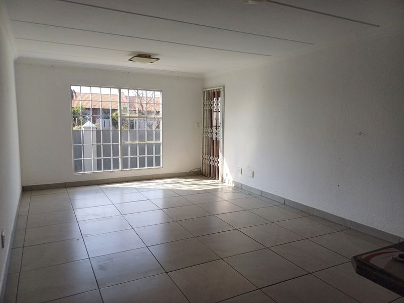 An empty, open-plan living area featuring neutral tiled flooring, a security gate, and large windows with burglar bars, offering a blank canvas for interior design.