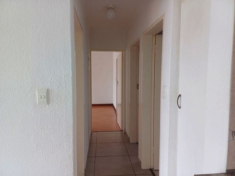A clean, neutral hallway featuring tiled flooring and multiple internal door access points, leading into an adjacent room with wood-look flooring.