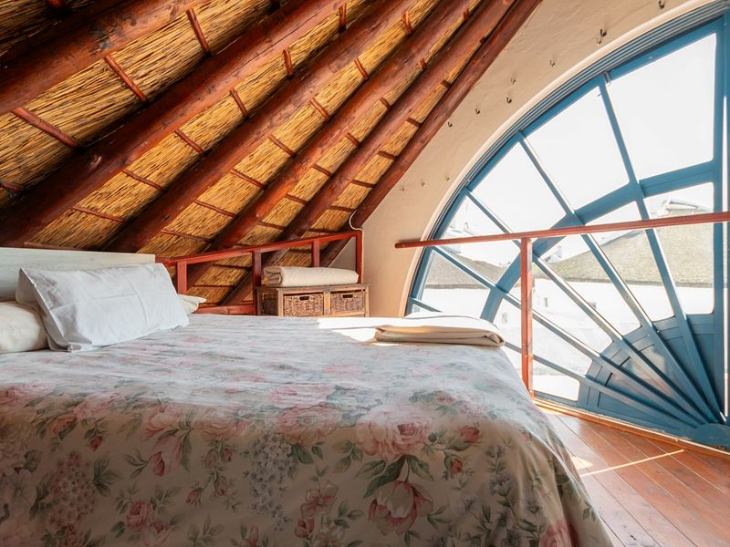 Unique loft-style bedroom featuring exposed timber rafters, a traditional thatched roof, and a large arched window providing ample natural light.
