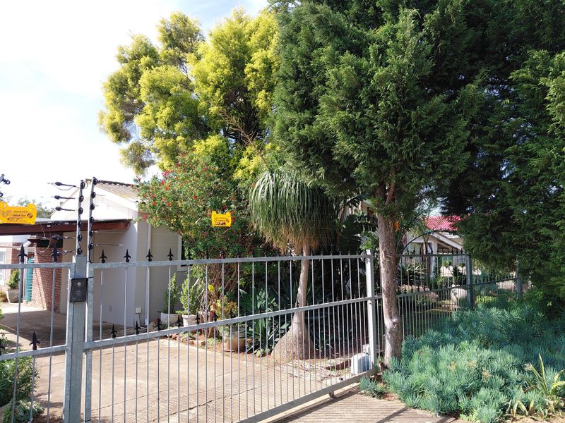 Front view of the property featuring a secure metal gate, electric fencing, and a concrete driveway surrounded by established garden landscaping.
