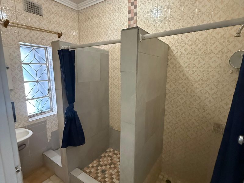 A functional bathroom featuring dual shower stalls with tiled finishes, a wall-mounted basin, and a window with security bars.