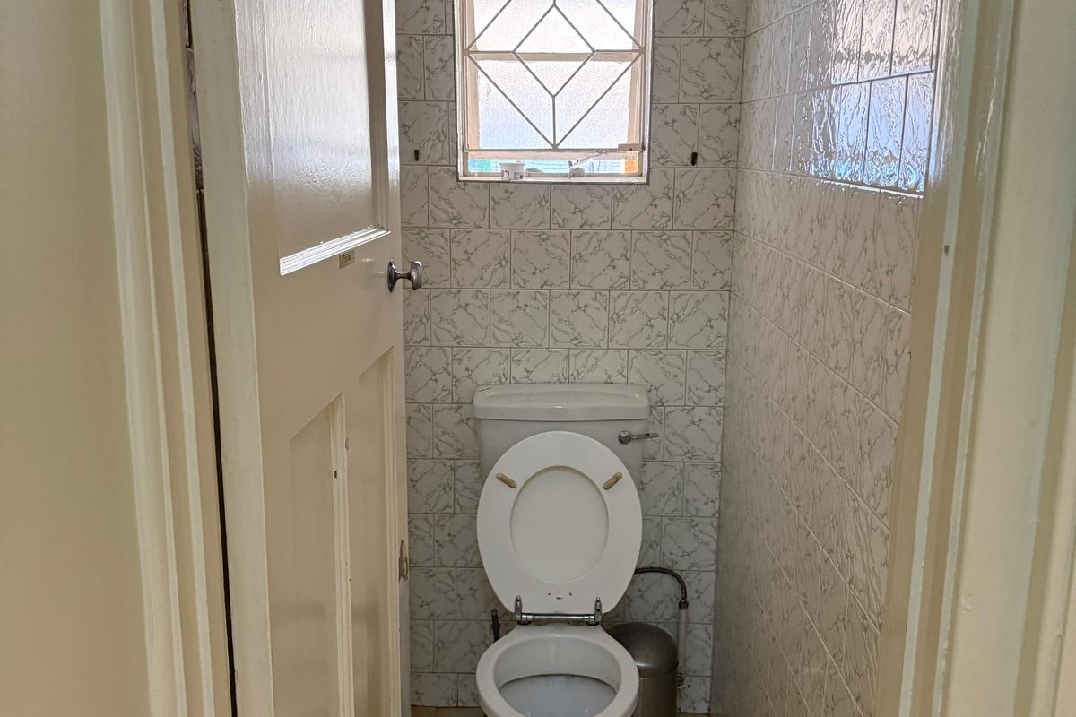 A compact, dated guest toilet featuring ceramic wall and floor tiles, a standard toilet suite, and a small window with security bars.