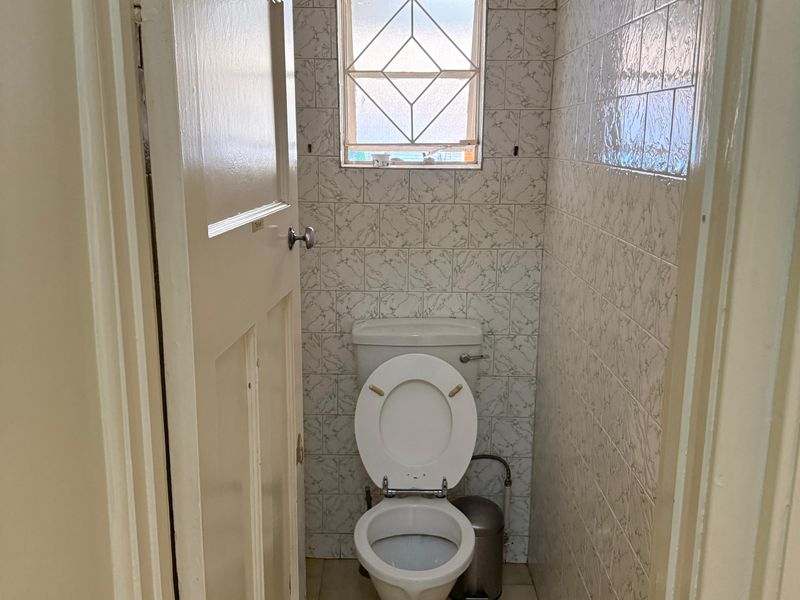 A compact, dated guest toilet featuring ceramic wall and floor tiles, a standard toilet suite, and a small window with security bars.