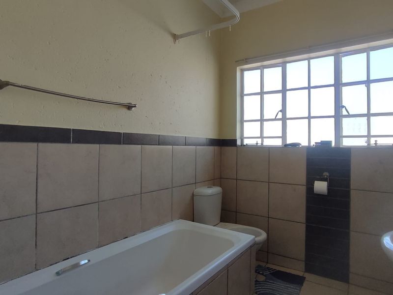 A functional bathroom featuring a bathtub, toilet, and basin with tiled walls and flooring, complemented by a window with security bars.
