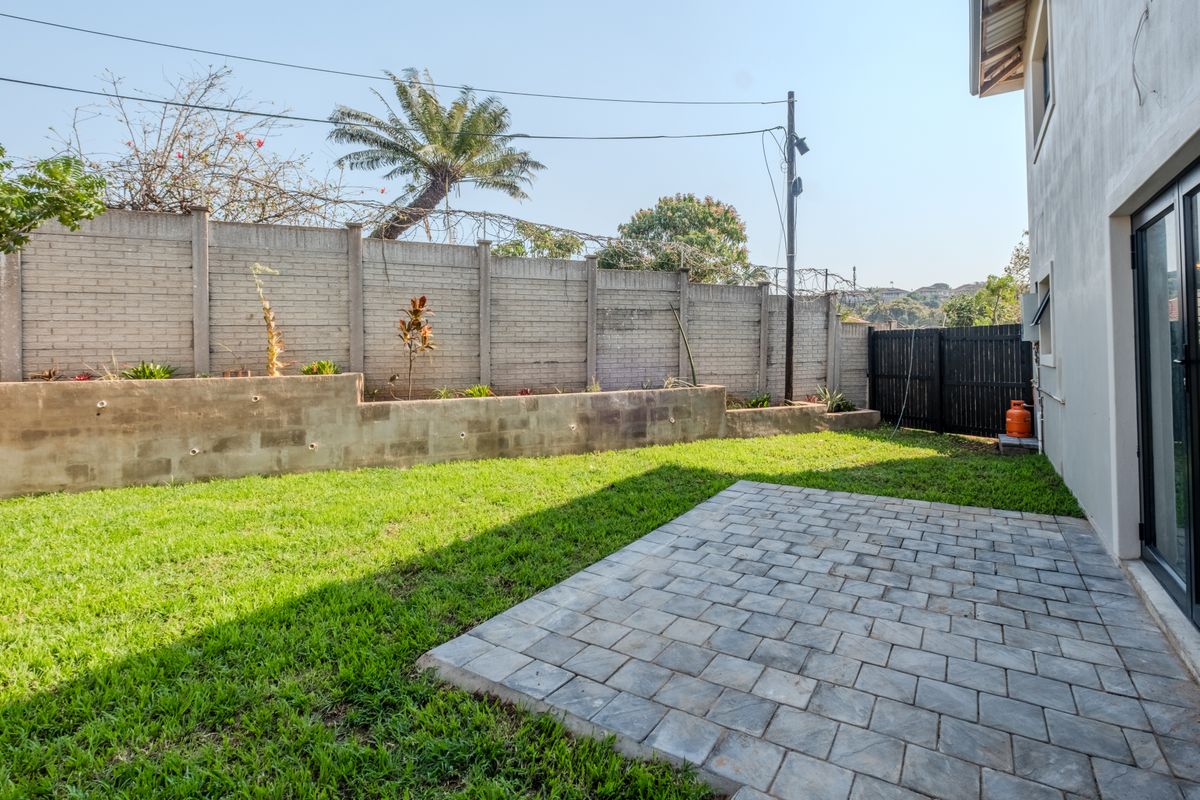 A private rear garden featuring a paved patio area, a grass lawn, and a concrete retaining wall with perimeter security fencing.