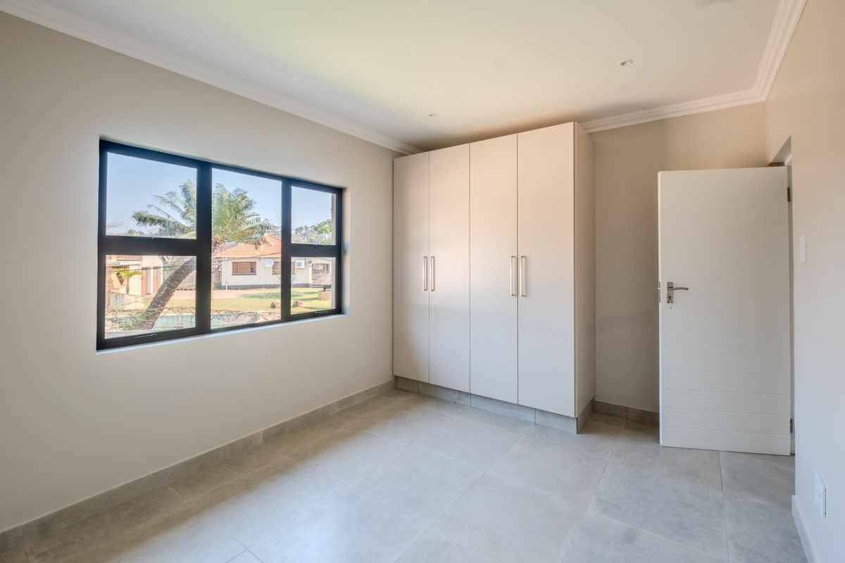 A bright, empty bedroom featuring neutral tiled flooring, built-in cupboards, and a large window providing natural light.