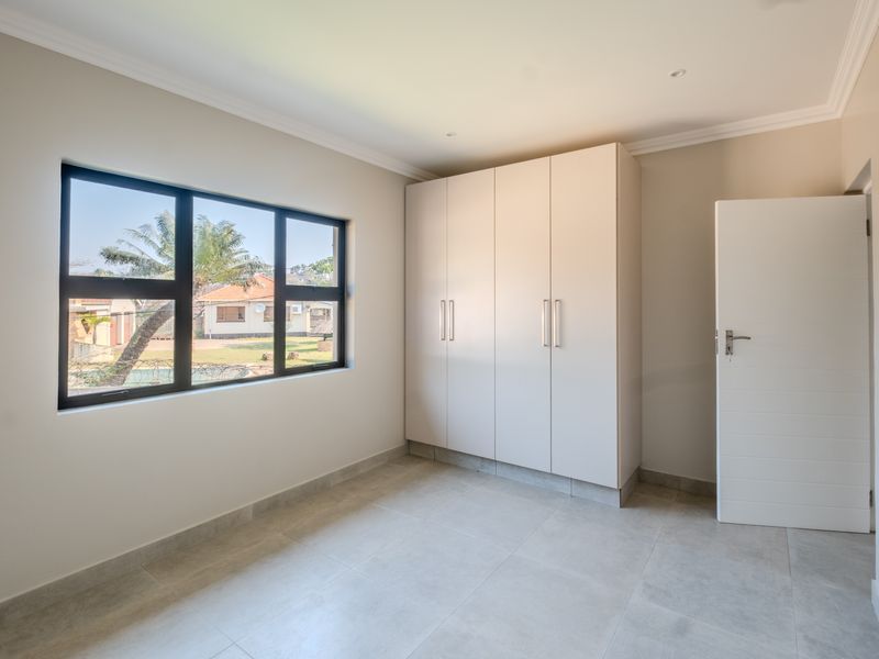 A bright, empty bedroom featuring neutral tiled flooring, built-in cupboards, and a large window providing natural light.