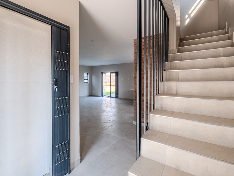 Bright entrance hall featuring a tiled staircase with metal balustrade, leading into an open-plan living area with exposed brick accents and neutral floor tiles.