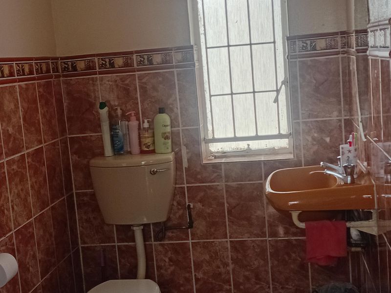 A compact bathroom featuring dated brown ceramic wall and floor tiles, a wall-mounted basin, and a standard toilet with security bars on the window.