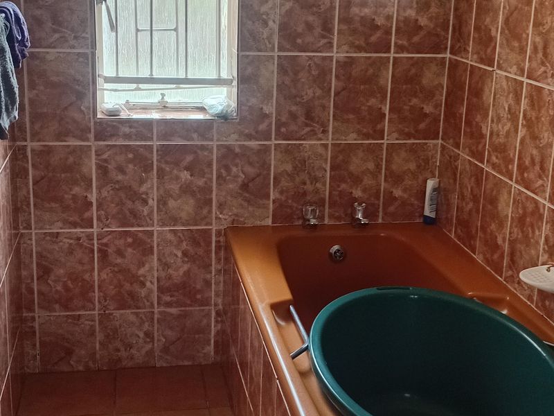 A dated bathroom featuring brown ceramic wall and floor tiles with a built-in bathtub and security bars on the window.