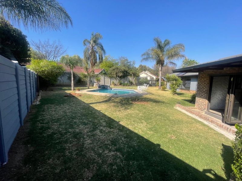 A spacious backyard featuring a swimming pool, established palm trees, and a brick-walled perimeter, complemented by a covered patio area.