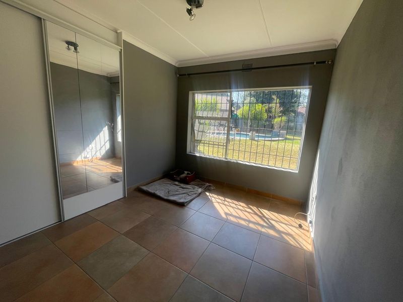 A bright, empty bedroom featuring tiled flooring, a large built-in mirrored wardrobe, and a window with security bars overlooking the garden.
