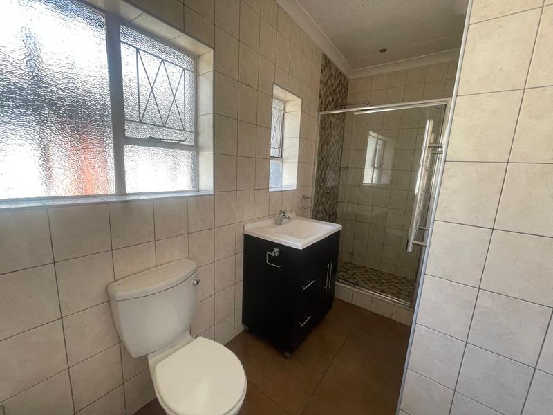A functional bathroom featuring a shower enclosure, vanity basin, and toilet with neutral wall and floor tiling.