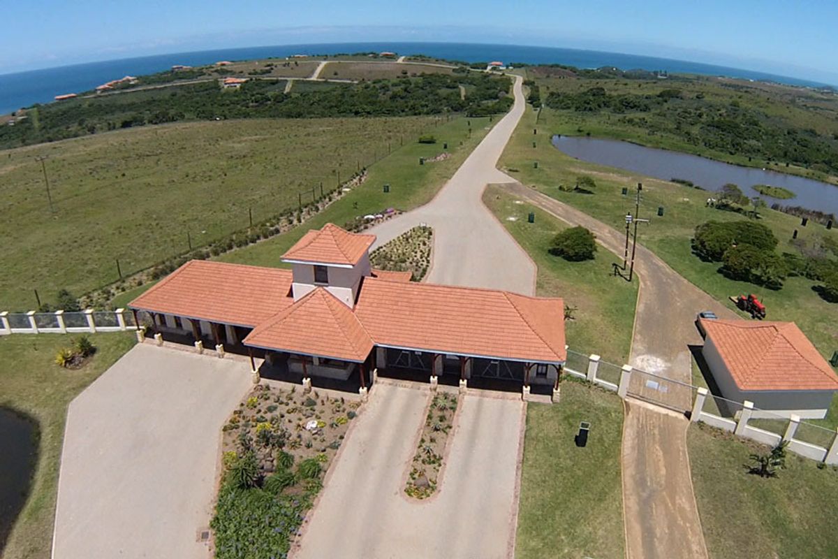 Aerial view of a residential estate entrance featuring a paved driveway, security guardhouse, and landscaped grounds with coastal surroundings.
