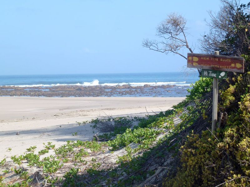 A scenic view of the coastline featuring sandy beach, rocky shoreline, and ocean waves, with a directional sign indicating high tide routes.