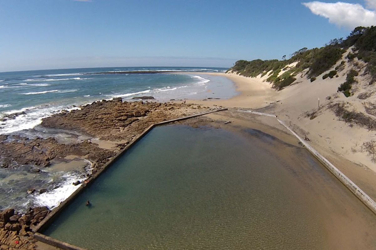 An aerial view of a natural coastal rock pool situated directly adjacent to the sandy beach and ocean shoreline.