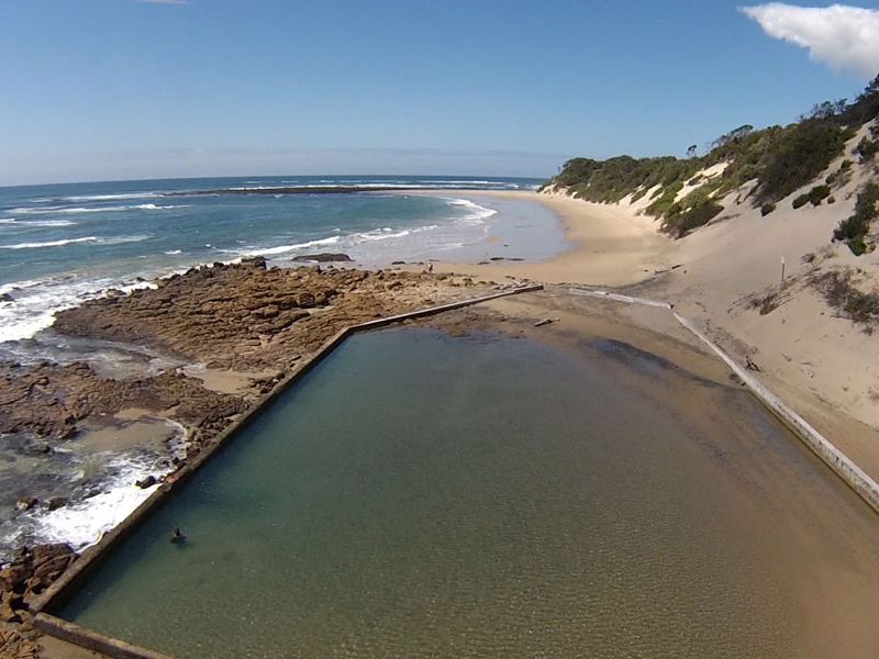 An aerial view of a natural coastal rock pool situated directly adjacent to the sandy beach and ocean shoreline.
