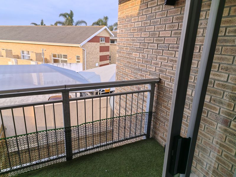 A compact balcony featuring brick walls, a metal balustrade, and artificial turf flooring, offering an elevated view of the surrounding townhouse complex.