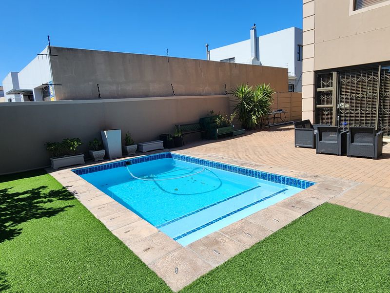 A private backyard featuring a swimming pool, paved patio area, and artificial lawn, enclosed by a perimeter wall for privacy.