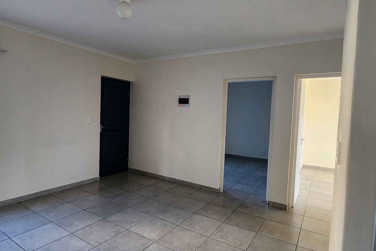 An empty, neutrally painted living area featuring tiled flooring, natural light from a window with security bars, and access to adjacent rooms.