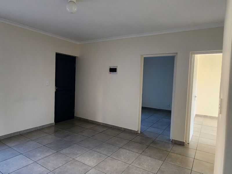 An empty, neutrally painted living area featuring tiled flooring, natural light from a window with security bars, and access to adjacent rooms.