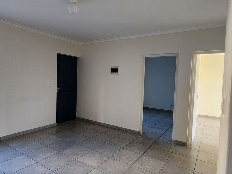 An empty living area featuring neutral tiled flooring, security bars on the window, and access to adjacent rooms.