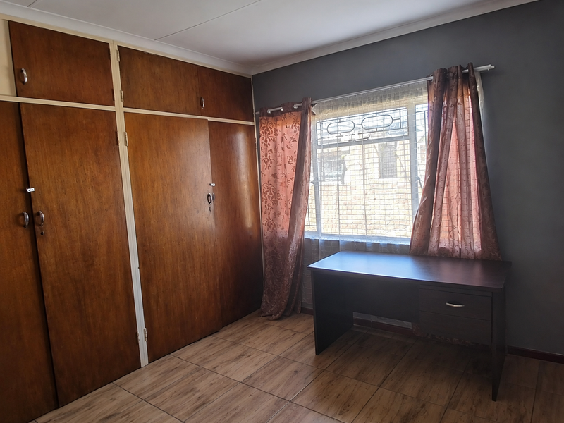 A bedroom featuring tiled flooring, built-in wooden cupboards, and a window with security bars, offering a functional and neutral space.