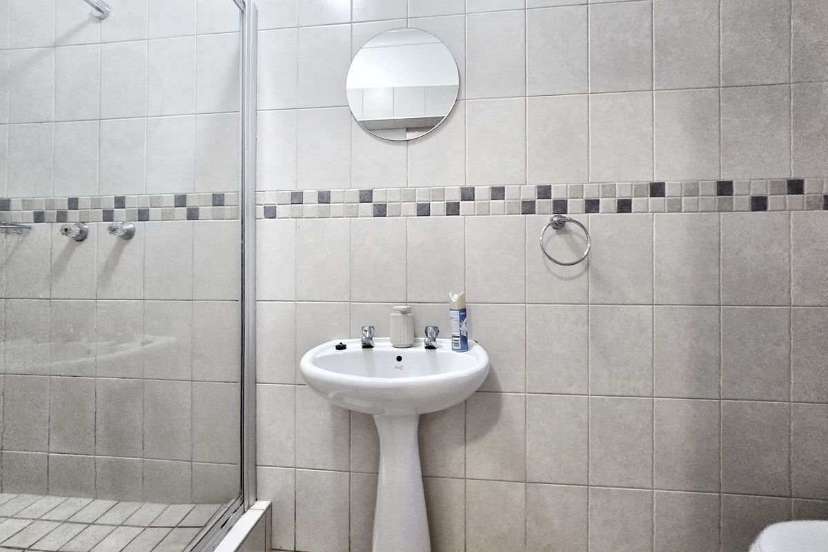A functional bathroom featuring a pedestal basin, a glass-enclosed shower, and neutral wall tiling with a decorative mosaic border.