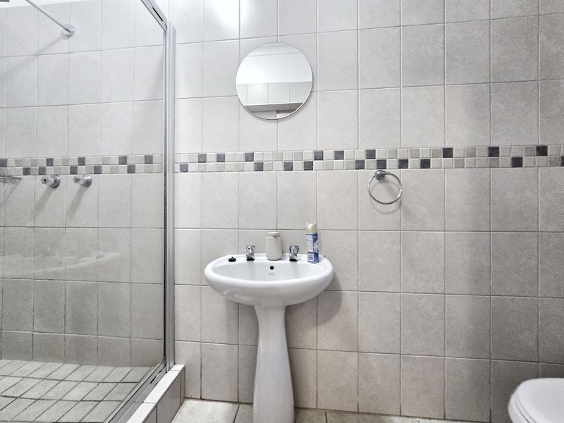 A functional bathroom featuring a pedestal basin, a glass-enclosed shower, and neutral wall tiling with a decorative mosaic border.