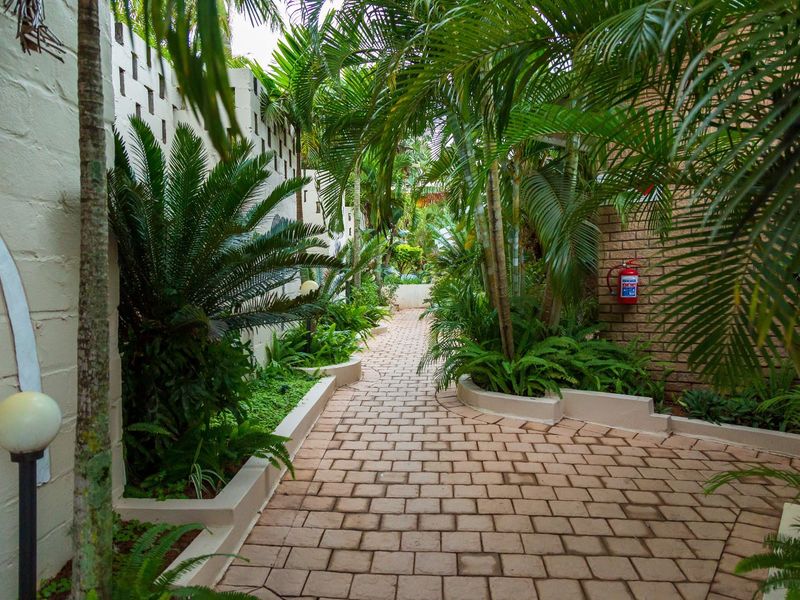 A well-maintained garden pathway featuring brick paving, lush tropical landscaping, raised garden beds, and secure boundary walls.