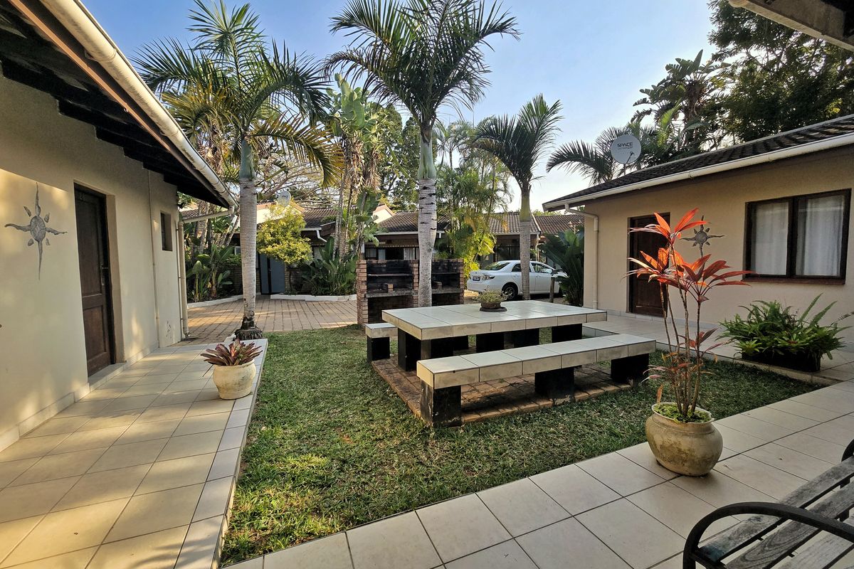 A well-maintained rear courtyard featuring a tiled patio, built-in brick braai, and a permanent tiled outdoor dining set surrounded by landscaped garden.