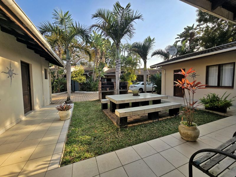A well-maintained rear courtyard featuring a tiled patio, built-in brick braai, and a permanent tiled outdoor dining set surrounded by landscaped garden.