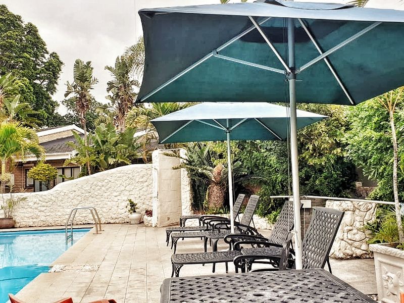 A well-maintained outdoor pool area featuring a paved deck, stone retaining wall, and established garden surroundings.