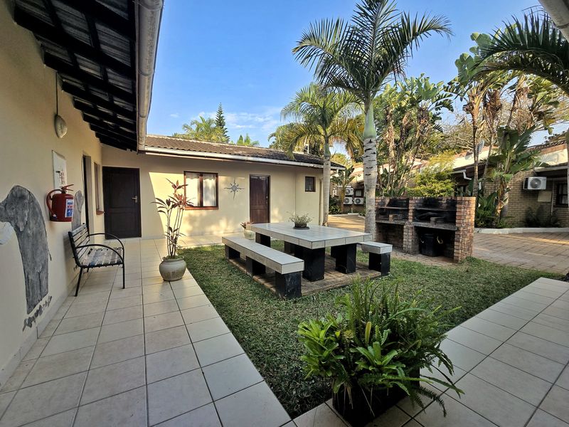 A well-maintained outdoor patio area featuring a built-in brick braai, tiled flooring, and a landscaped garden setting.