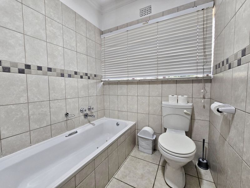 A functional bathroom featuring a built-in bathtub, toilet, and neutral tiled finishes with a large window covered by venetian blinds.