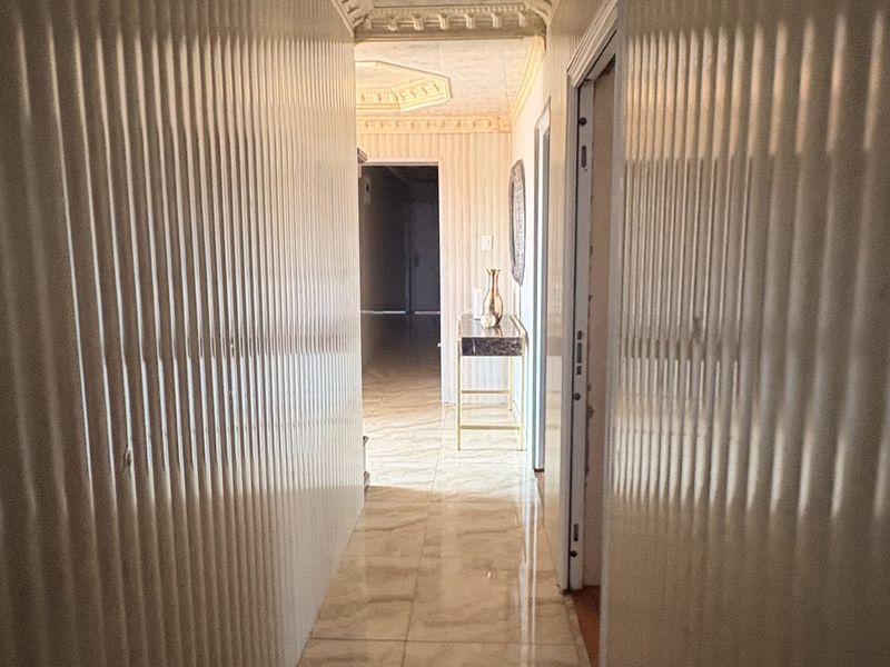 A long, narrow hallway featuring polished tiled flooring, textured wall paneling, and decorative ceiling molding, leading towards an open living space.