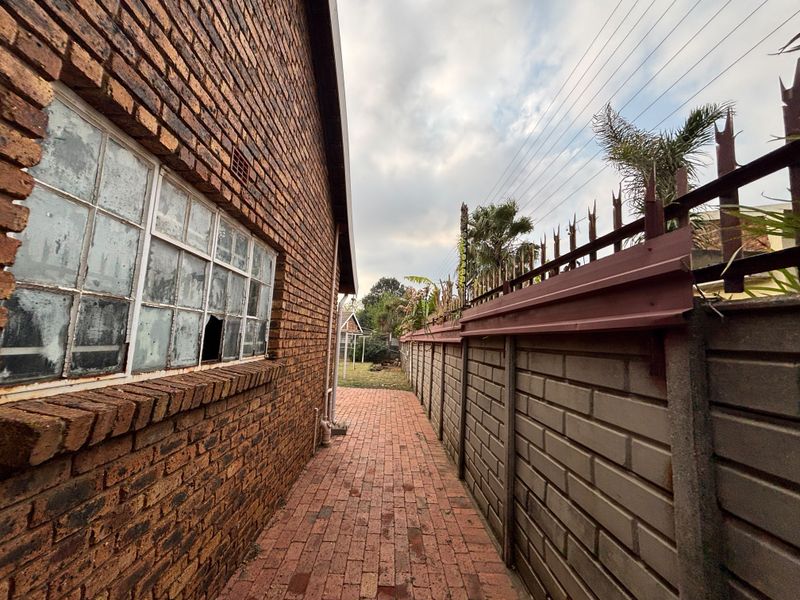 A side exterior view showing a face brick house wall with a damaged window, a brick paved walkway, and a high boundary wall with security spikes.