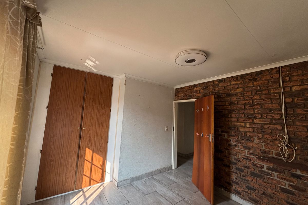 A bedroom featuring an exposed brick wall, built-in wooden cupboards, and tiled flooring. The room is vacant and requires minor cosmetic updates.