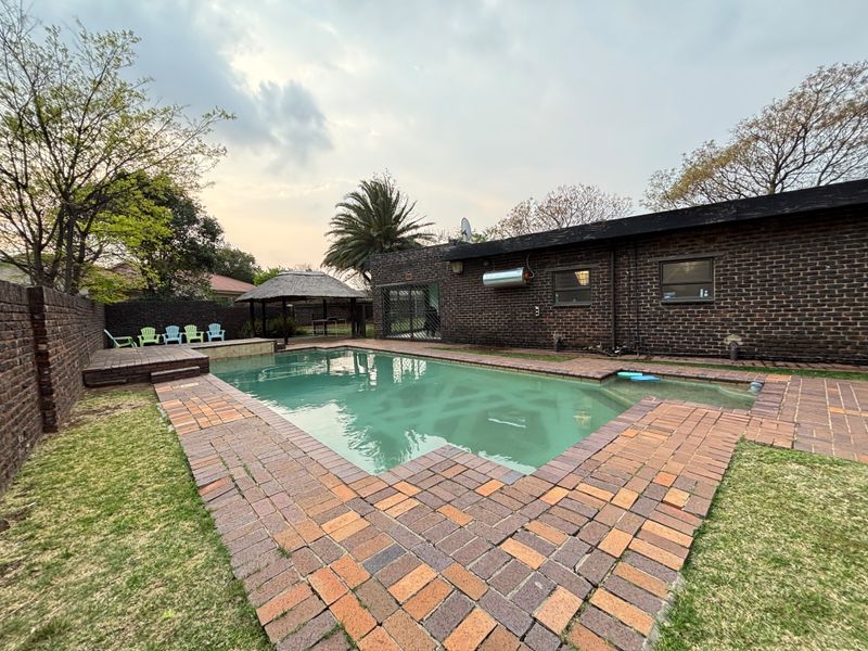 Outdoor pool area featuring a swimming pool, brick paving, and a thatched gazebo set within a private garden with brick boundary walls.