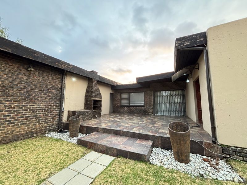A paved outdoor patio area featuring a built-in brick braai, accessible via a security gate, set against a brick and plastered exterior wall.