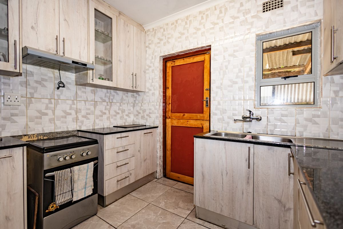 Functional kitchen featuring light wood-grain cabinetry, dark countertops, tiled splashbacks, and a stainless steel sink with a dedicated exterior access door.