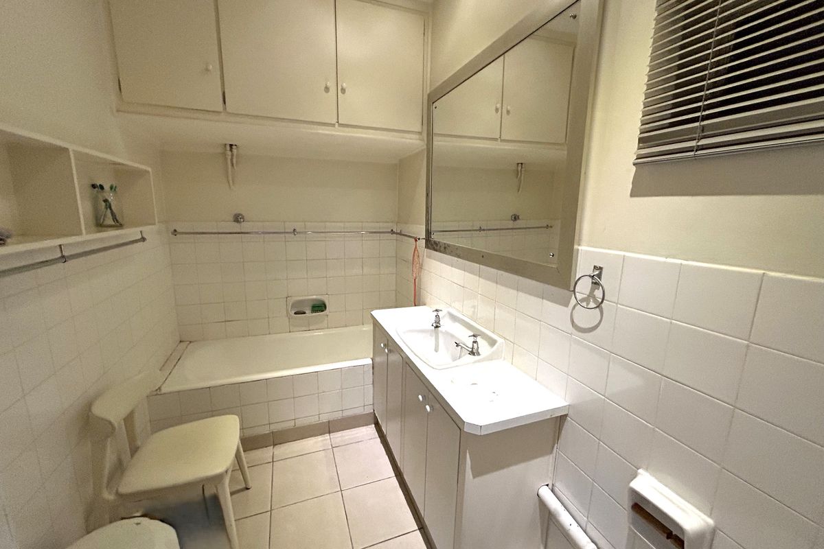 A functional bathroom featuring a tiled bathtub, built-in vanity with storage, wall-mounted mirror, and overhead cabinetry, offering a classic layout with potential for modernization.