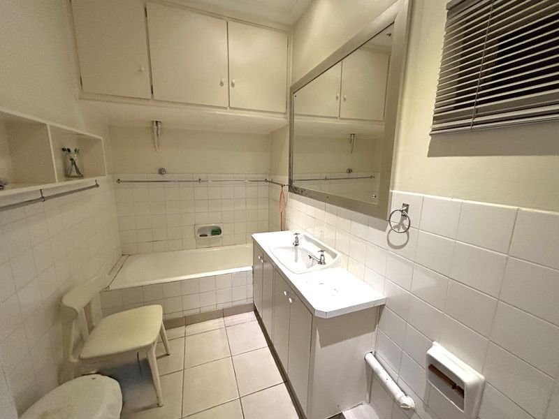A functional bathroom featuring a tiled bathtub, built-in vanity with storage, wall-mounted mirror, and overhead cabinetry, offering a classic layout with potential for modernization.