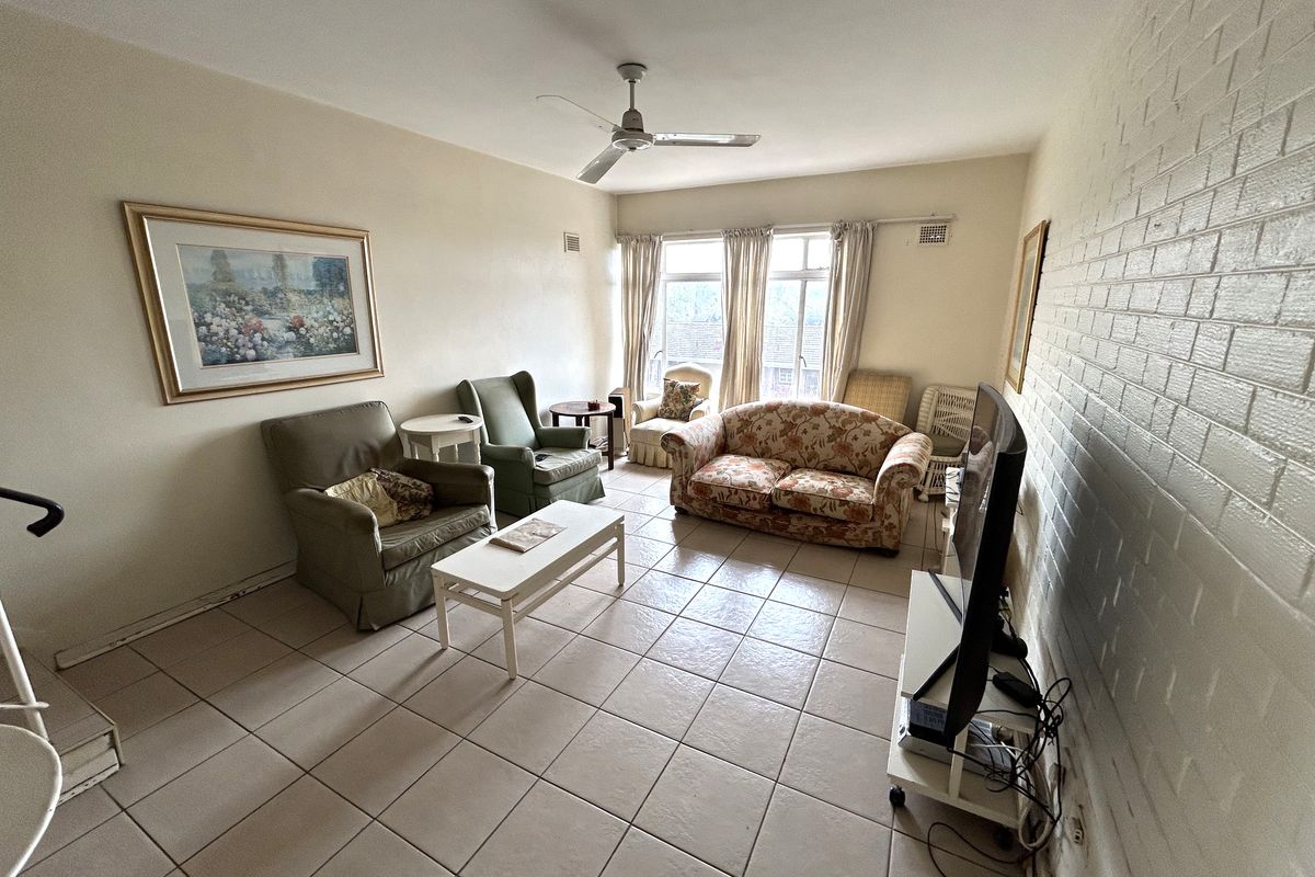 A spacious living room featuring tiled flooring, a ceiling fan, and a painted brick feature wall with natural light from a large window.
