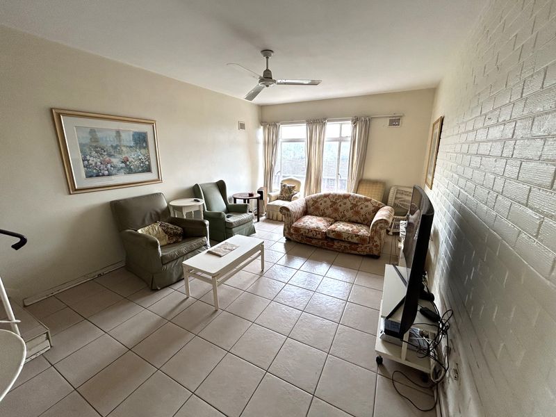 A spacious living room featuring tiled flooring, a ceiling fan, and a painted brick feature wall with natural light from a large window.
