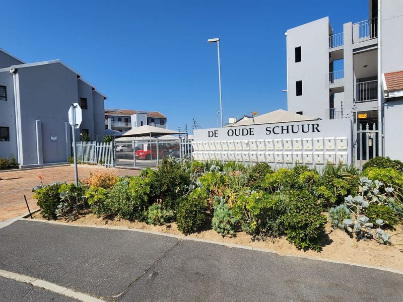 Front entrance view of De Oude Schuur apartment complex featuring a paved driveway, communal post boxes, and well-maintained landscaped garden areas.